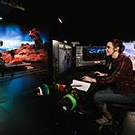 A woman is sitting at a desk in front of several monitors. She is in a virtual production studio that has been designed to look similar to Mars.